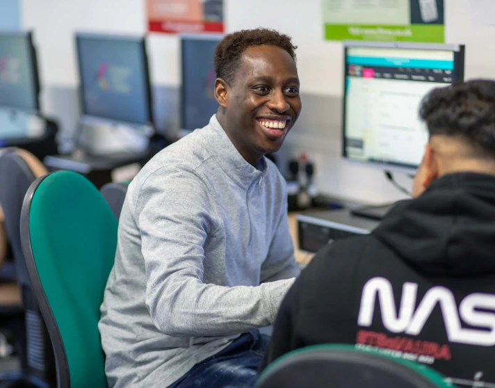 Two students working together at a computer in a business and administration course, one smiling while engaged in discussion. Two students working together at a computer in a business and administration course, one smiling while engaged in discussion.
